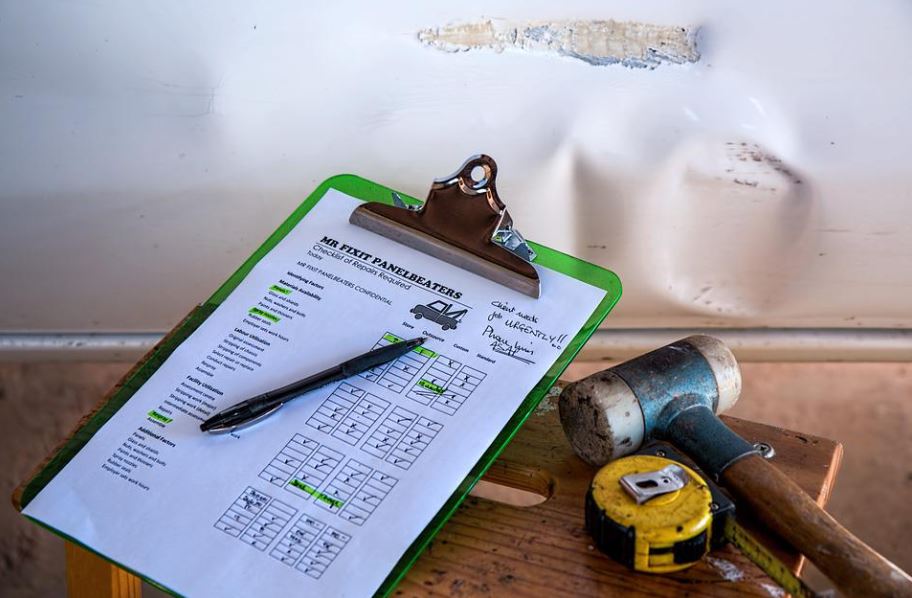 a damage repair checklist beside a tape measure and a mallet, and a damaged car at the back