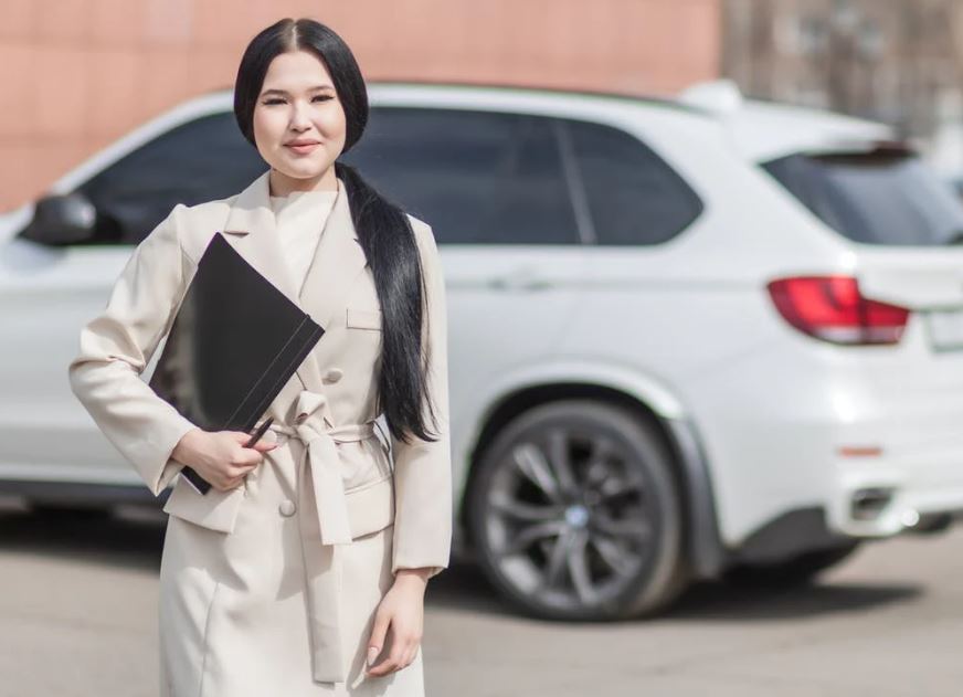 a woman in beige corporate clothes holding a black folder and a white car behind her