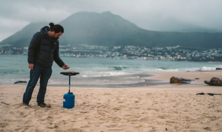 a man cooking at the beach using a portable gas cylinder