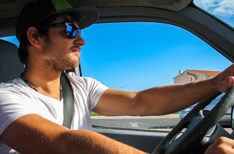 man in white t-shirt, sunglasses, and cap wearing his seatbelt while driving