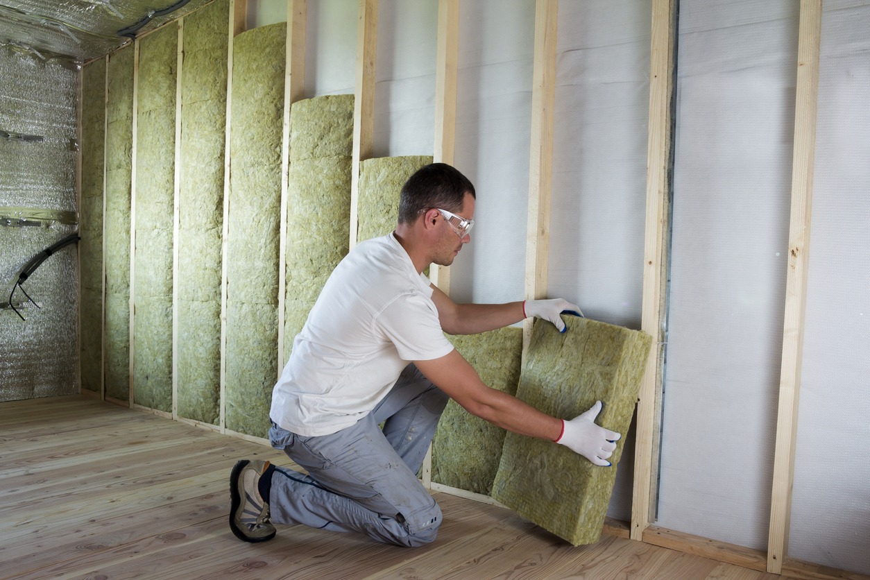Worker in protective goggles and respirator insulating rock wool insulation in wooden frame